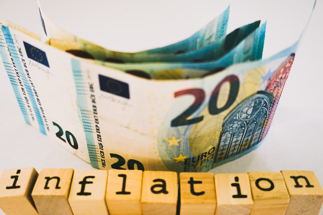 A thought-provoking still life featuring a rolled 20 euro banknote with its vibrant colors and security details prominently displayed. Below it, rustic wooden blocks spell out the word inflation, creating a striking visual metaphor for the economic forces that impact currency value. The clean white background and soft lighting draw attention to the contrast between the crisp, modern banknote and the raw texture of the blocks—ideal for discussions on finance, economics, or educational content about monetary policy.